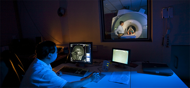 Photo shows a researcher looking at a computer screen showing a scan of a human brain. Through a lab window, a child lays in the center of a large MRI machine, accompanied by another researcher.
