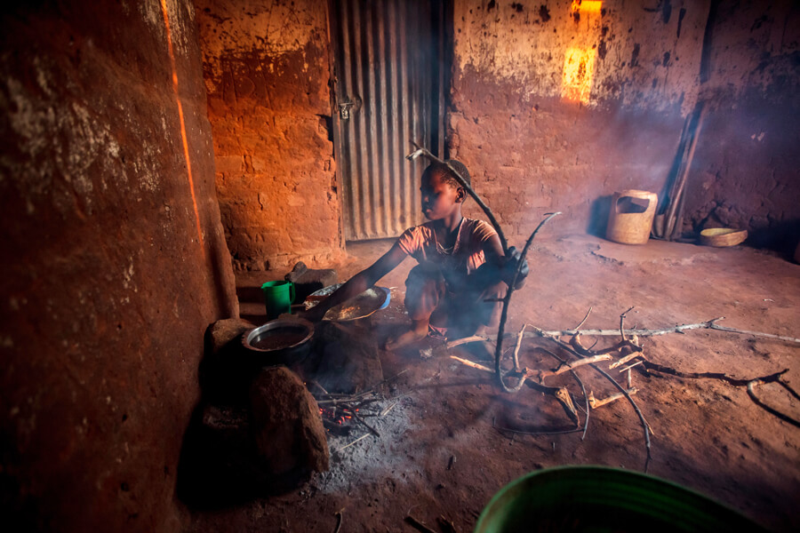 A young girl squats over a stove made of stone and fueled by firewood. Smoke rises from the indoor stove, which sits on the dirt floor of an unfurnished room.