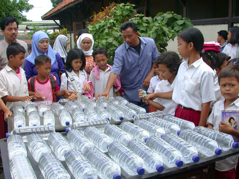 In a community in Indonesia, a group of people in a yard are gathered around two tables, where about four dozen clear, plastic bottles of water lay on their sides.