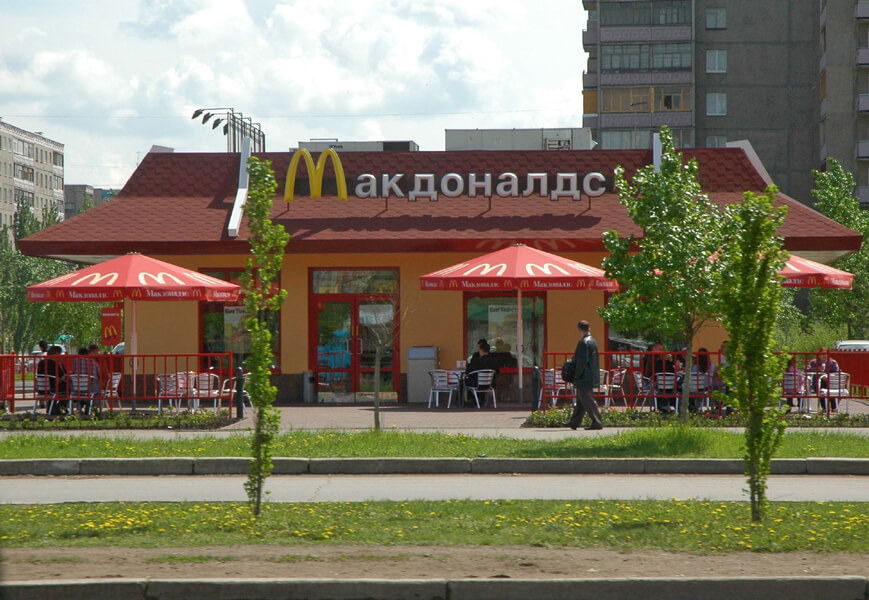 Photograph of a McDonald’s restaurant with Russian signage. People are seated outside the restaurant under red umbrellas that are decorated with small golden arches.