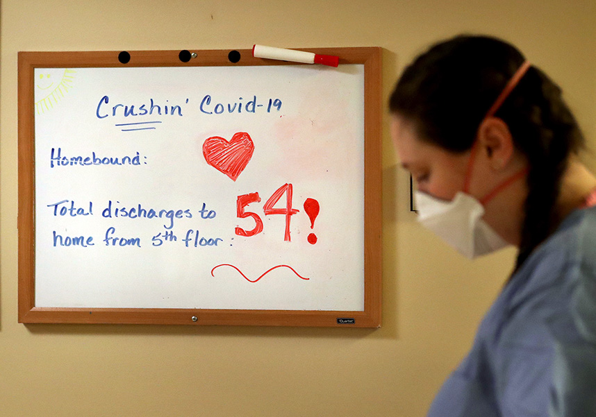 Photograph of a whiteboard that says “Crushin’ Covid-19. Homebound: Total discharges to home from 5th floor: 54!” A healthcare worker in blue scrubs and white mask stands next to the board.