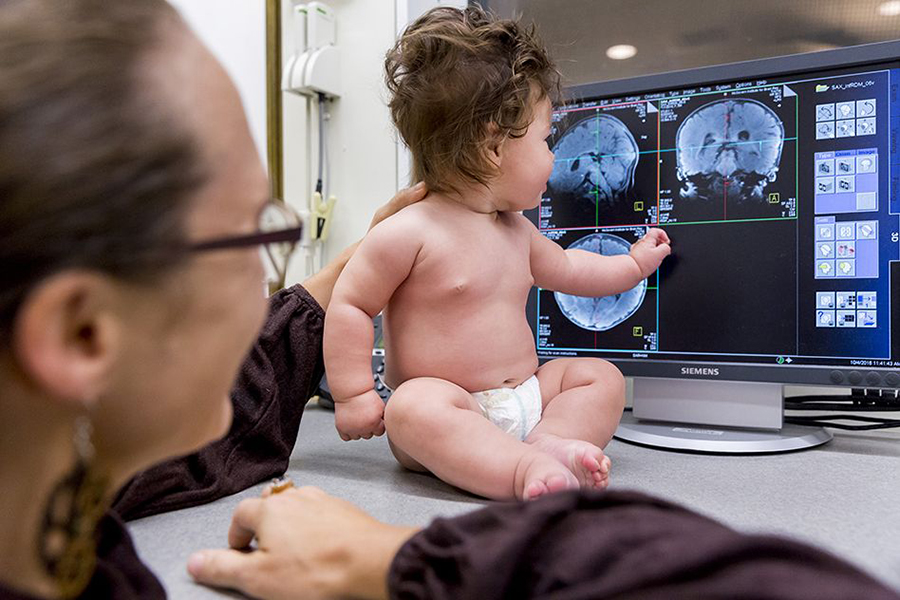 A baby wearing only a diaper sitting on a desk looks at MRI scans of an infant brain displayed on a computer while a woman supports the baby’s back.