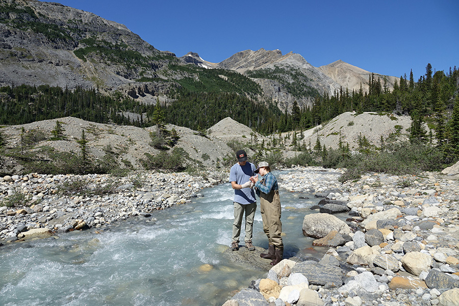 Photograph of two people standing on a large rock in a stream, examining a sample. Stark mountains surround them.