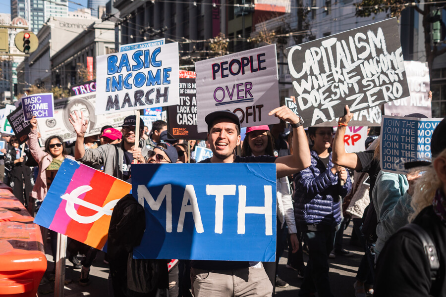 Photograph of a crowd of demonstrators marching down a city street. They are holding banners that say, among other things, “Basic income march,” “People over profit” and “Economic justice for all.”