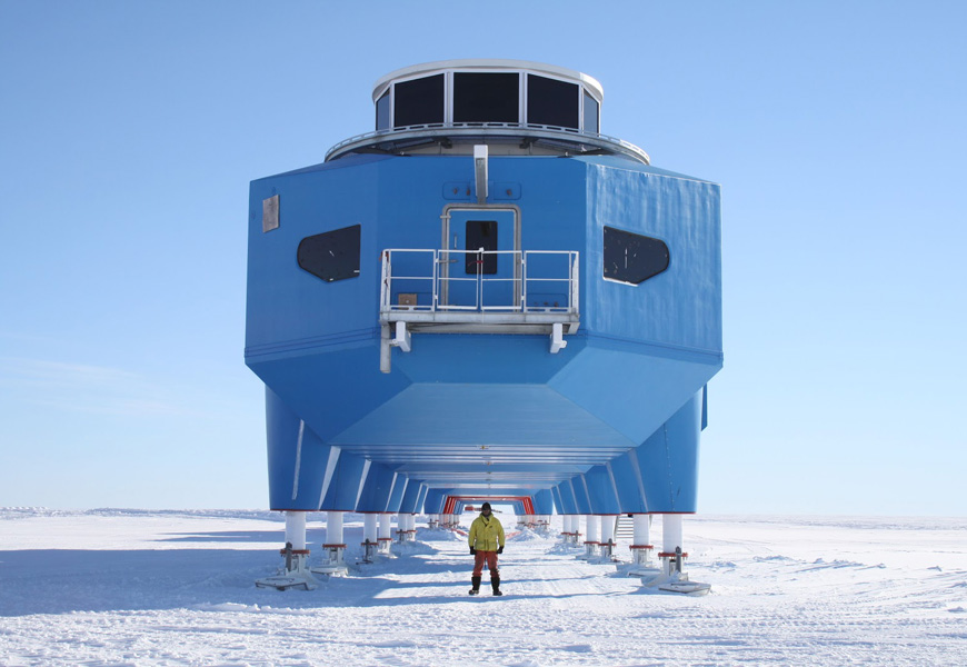 Photograph of a man in warm, protective clothing standing outdoors in flat, snow-covered terrain. He is in front of a strange blue building on stilts. The building is long but is viewed from head-on.