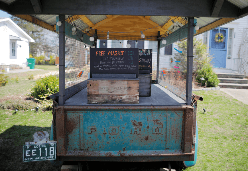 A covered container stands on a grass lawn with a house in the background. Inside the container is a sign that says, “free masks.”