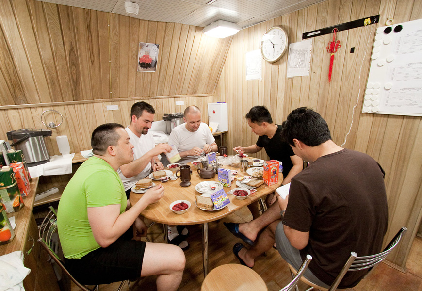 Five men sit around a table eating bread, fruit and other food in a small room.