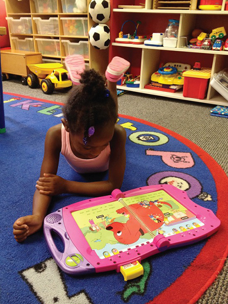 A young child sits on a mat looking at a story in a PowerTouch console. There are shelves with toys in the background.