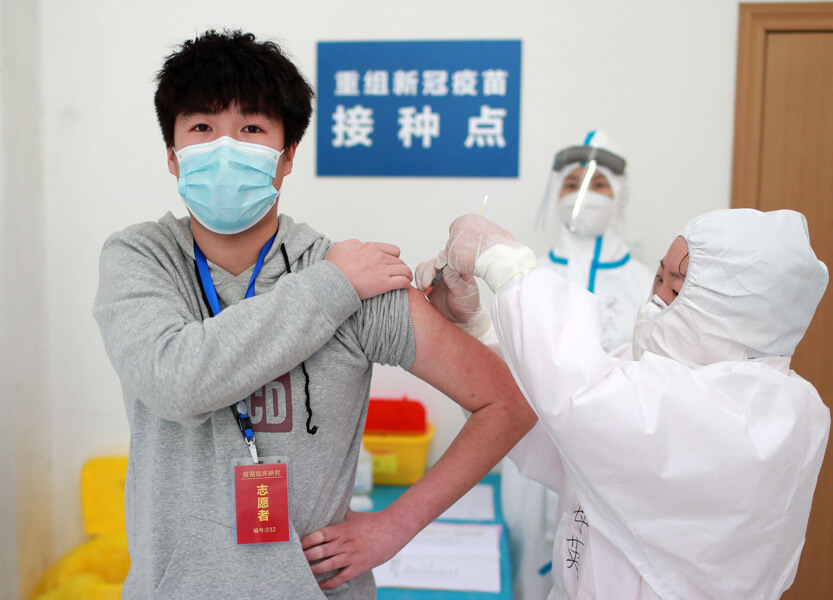A man in a grey sweatshirt wearing a surgical mask has rolled up his left sleeve and is receiving an injection in his upper arm. It is being administered by a person heavily suited up in white protective gear. Another suited-up person stands in the background wearing a clear visor over their face.