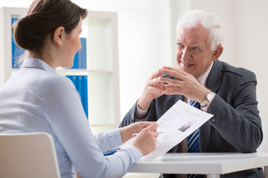 Photo shows an elderly man in a suit being interviewed by a young woman, who holds a copy of his resume.