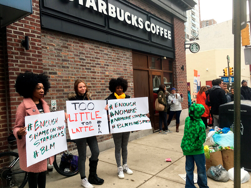 This 2018 news photo shows three women demonstrating with signs outside of a Starbucks café on a crowded street corner in Philadelphia.