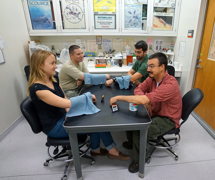 Photograph shows four individuals sitting around a table. Each person has one arm resting on a large white paper cup containing mosquitoes.