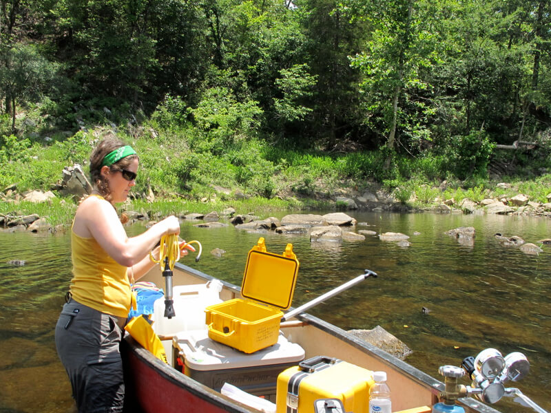 Photograph shows a researcher holding water-sampling equipment as she stands in a shallow river alongside a canoe filled with various pieces of lab equipment.