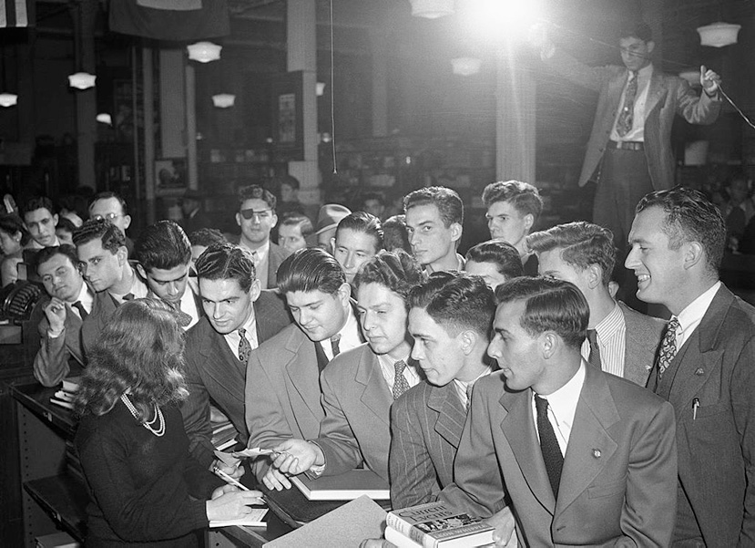 Historic photo from 1945 shows a crowd of recent military veterans gathered to receive school supplies and books as part of the G.I. Bill. A woman behind a bookcase takes orders. 