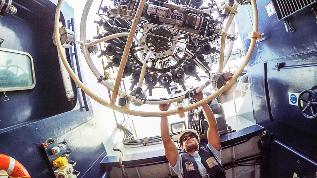 In a photo taken during the Schmidt Ocean Institute’s “Voyage to the White Shark Café” expedition last year, boatswain Mick Utley is holding part of the CTD rosette, an instrument with black bottles lined up in a circle.
