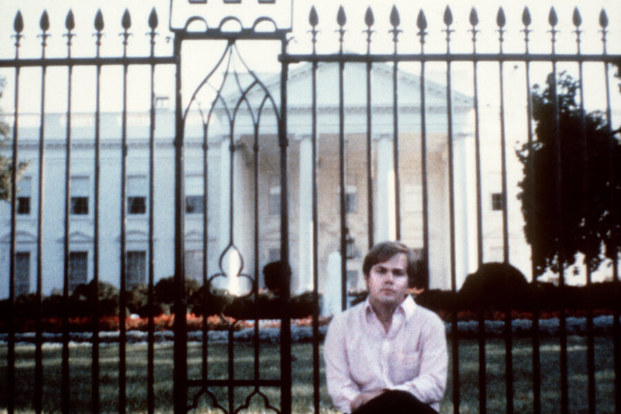 Photograph of John W. Hinckley Jr. wearing a light shirt and dark pants. Behind him are railings, and the White House is in view behind them.