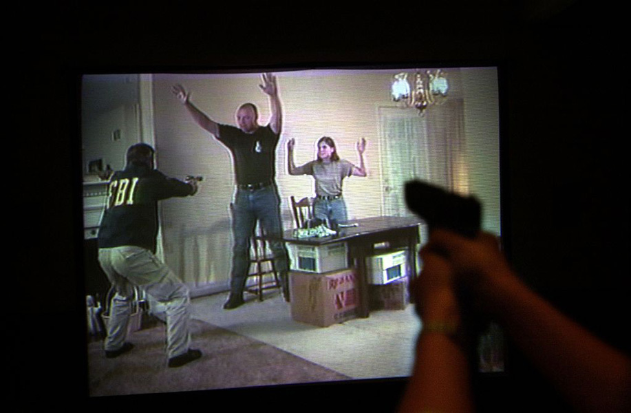 Photograph of a screen showing an image of an FBI agent aiming a gun at a Black man and white woman, both with their hands in the air. Outside of the screen, a person’s hands are seen pointing a gun at the image.