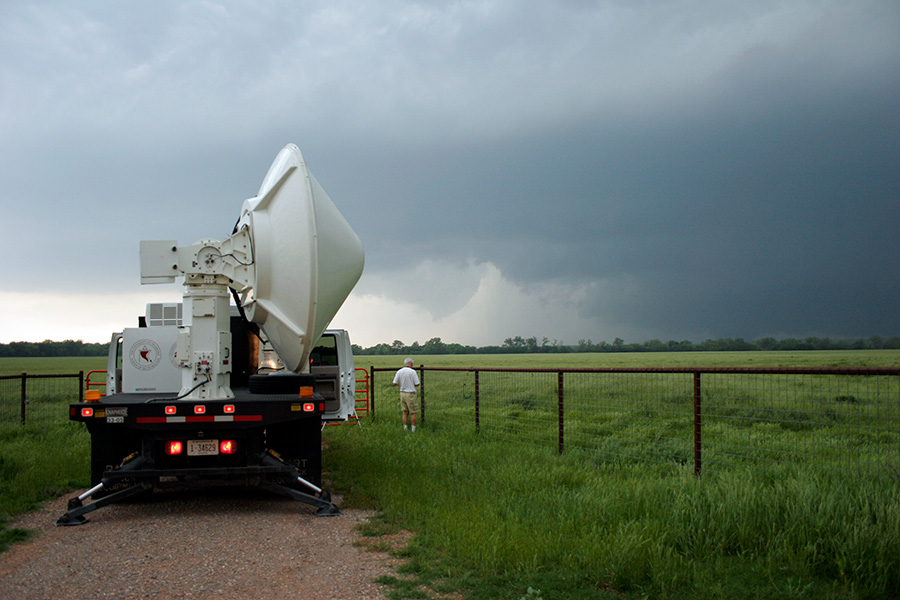 A truck seen from the back with a giant radar contraption in its bed, a person stands nearby looking at tall clouds spawning a tornado in the distance
