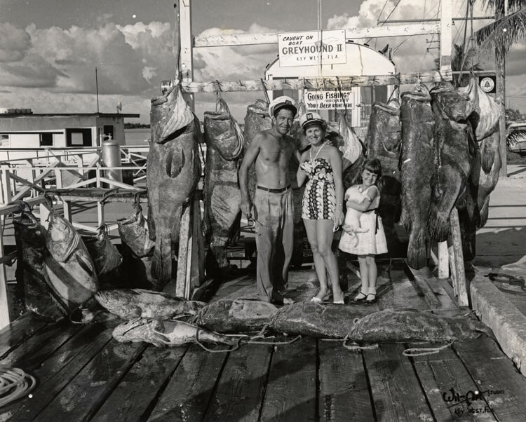 Photograph of a shirtless man in a captain’s hat standing next to a woman in a bathing suit and a little girl holding a pet dog. A haul of large fish is displayed on the ground in front of them and strung up behind them.