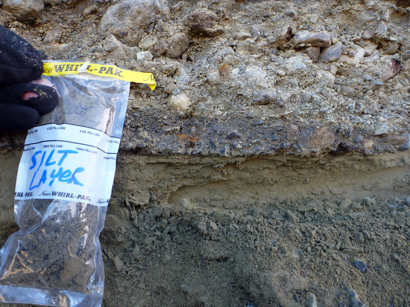 Photo shows a close-up of dirt layers, including rocks on top and fine sediments forming a narrow band below, with another layer of larger sediments below that. A sample collection bag is taped to the rocks.