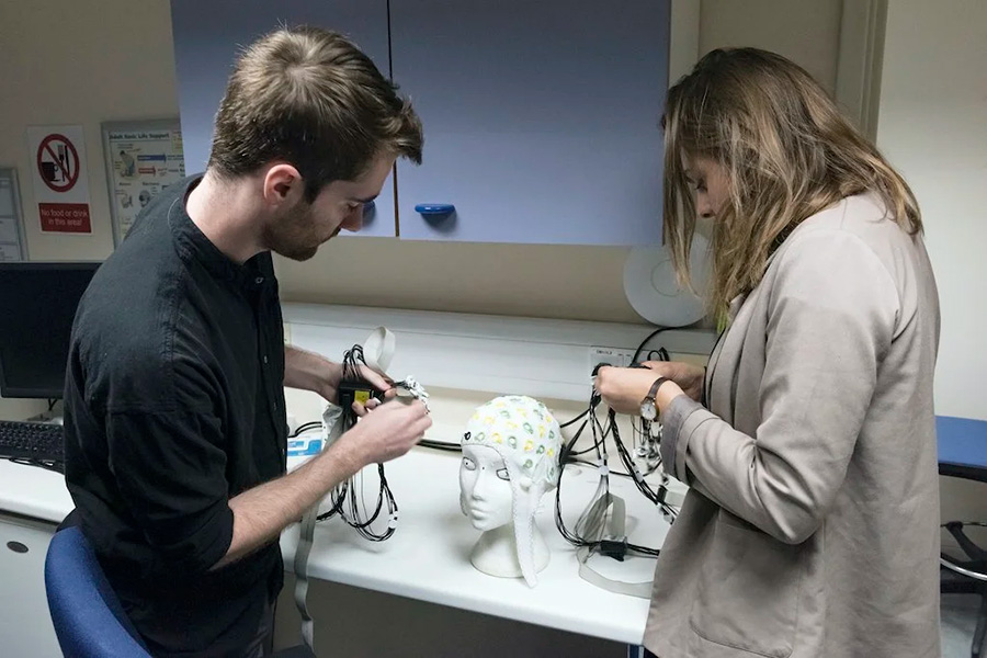 EEG cap Photograph of two individuals working with electronic equipment on a lab bench. They are leaning over a model of a human head upon which a cap with electrodes is placed.