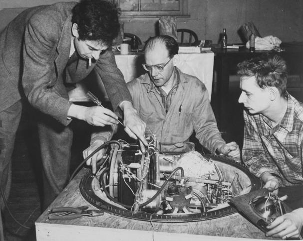 Old photo shows man in suit standing, cigarette in mouth, while adjusting machinery on an instrument while a seated technician and younger man look on.