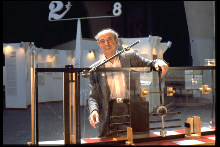 Color photo shows an older man standing behind a contraption on a table.