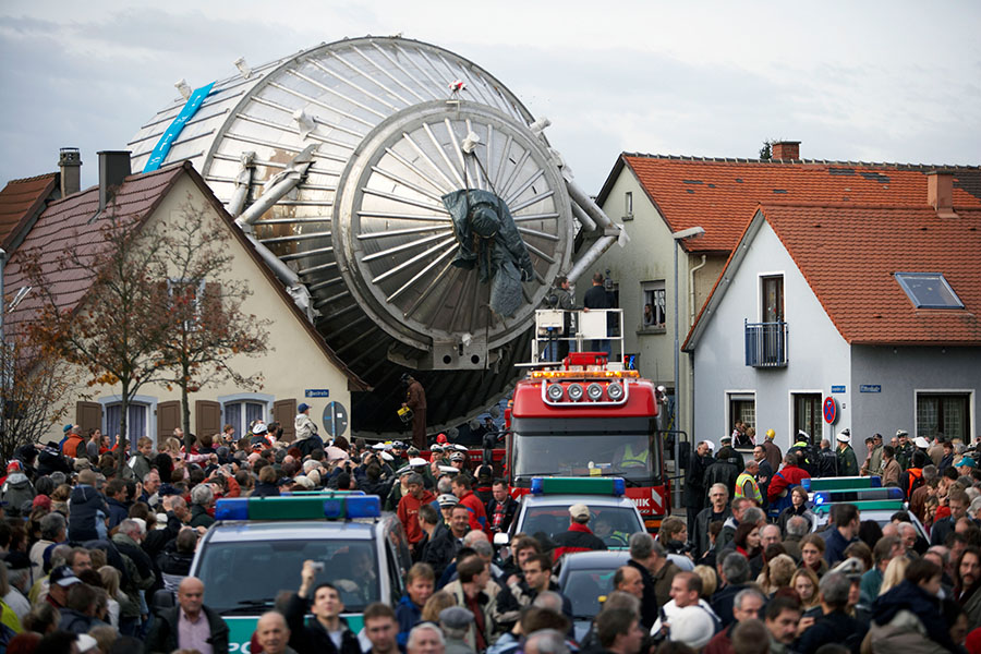KATRIN in transit Photo shows the transport of the massive, metallic KATRIN spectrometer down a narrow street, overhanging the houses on either side, in the center of Leopoldshafen, Germany, with people gathered for the event.