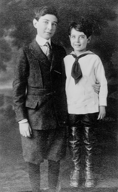 An old-fashioned black-and-white studio photo shows two boys, one younger in a sailor suit and long boots, the older taller one in a tweed suit.