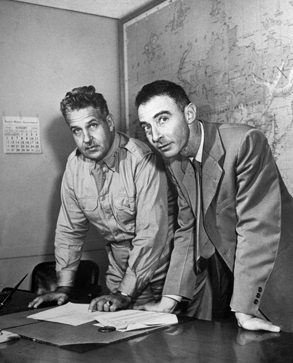 A black-and-white photo of two men, one in a military uniform and the other in a suit, stand before a desk, with a map behind them.