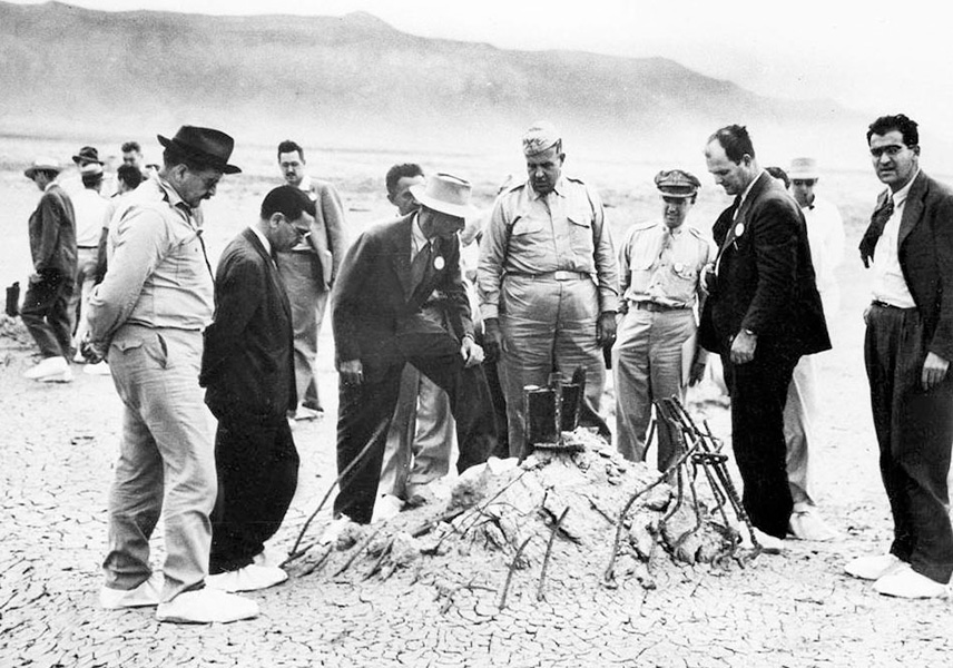 A black and white photo shows a small group of men in suits and uniforms surrounding a mound of dirt with bent steel rebar poking up through it, against a desert backdrop.